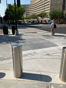 A picture of the intersection at D and 7th Streets. There are bollards in the foreground, and in the background, a person about to cross the street using a crosswalk that goes up and to the left.